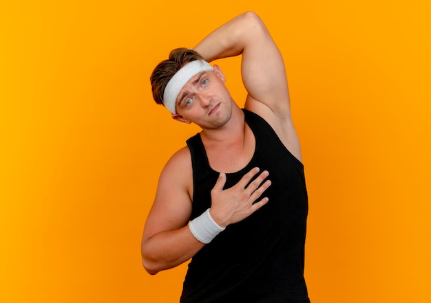 Foto grátis homem jovem bonito e confiante desportivo usando bandana e pulseiras, colocando as mãos no peito e atrás da cabeça, isolado em laranja com espaço de cópia