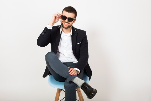 Homem jovem bonito elegante hipster em jaqueta preta, estilo empresarial, camisa branca, isolado, sentado relaxado na cadeira do escritório, falando no smartphone, sorrindo, feliz, positivo, óculos de sol