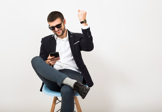 Homem jovem bonito elegante hipster em jaqueta preta, estilo empresarial, camisa branca, isolado, sentado relaxado na cadeira do escritório, usando smartphone, sorrindo, feliz, positivo, óculos de sol, gesto, emocional