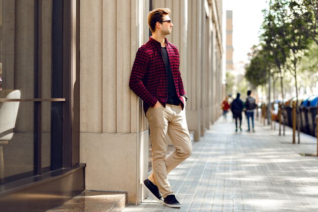 Homem jovem bonito hippie posando na rua europeia, ensolarados tons quentes cores, roupas da moda casuais, clima de viagem.