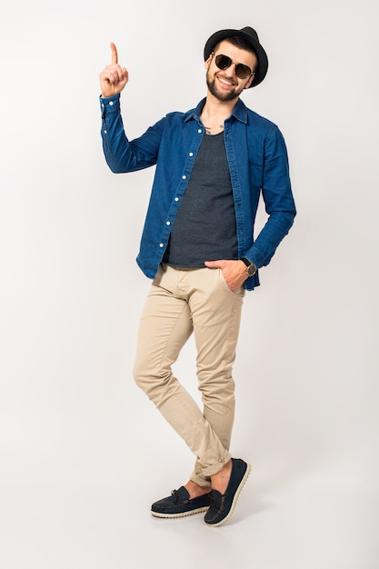 Homem jovem bonito hippie, roupa de estilo moderno, camisa jeans, calças, óculos de sol, chapéu, isolado, pulando, alegre, dedo apontando