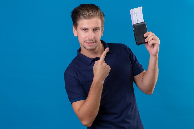 Foto grátis homem jovem bonito viajante segurando bilhetes aéreos, apontando o dedo para eles feliz e positivo, olhando para a câmera com sorriso confiante no rosto em pé sobre fundo azul