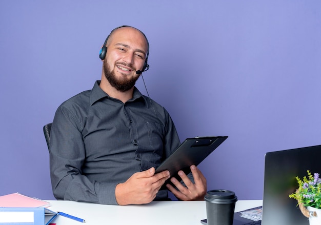 Homem jovem careca de call center usando fone de ouvido, sentado na mesa com ferramentas de trabalho isoladas em um fundo roxo