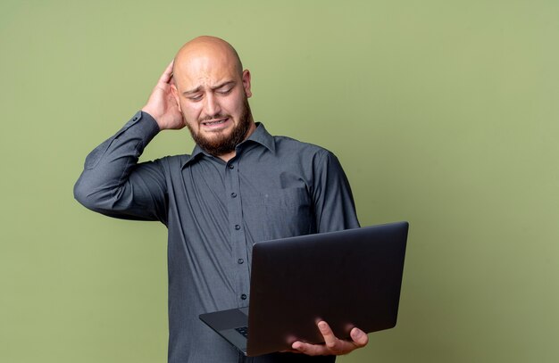 Homem jovem careca irritado de call center segurando e olhando para um laptop com a mão na cabeça isolada em verde oliva com espaço de cópia