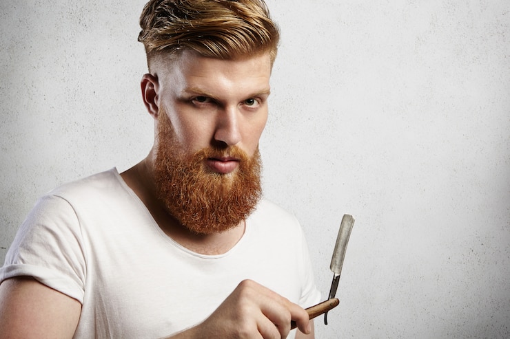 Foto grátis homem jovem caucasiano com aspecto de hipster em camiseta branca tentando decidir se raspa sua longa barba ruiva ou não. cara elegante segurando navalha com expressão facial séria e aparência.