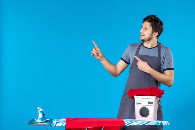 Homem jovem com tábua de passar roupa em um fundo azul homem jovem cor de ferro limpeza homem máquina de lavar roupa trabalho doméstico