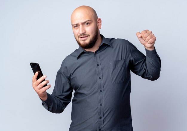 Homem jovem confiante e careca de call center segurando um telefone celular e cerrando o punho isolado no branco