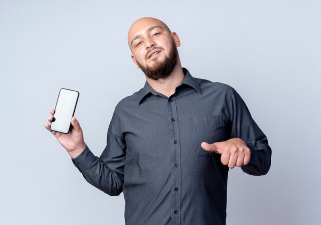 Homem jovem confiante e careca no call center, mostrando o celular e apontando para ele, isolado no branco