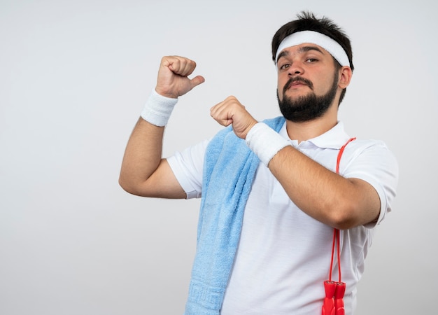 Homem jovem confiante e esportivo usando bandana e pulseira apontando para trás com uma toalha na corda de pular no ombro isolado na parede branca