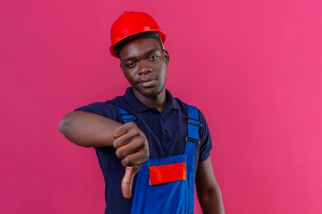 Homem jovem construtor afro-americano descontente com uniforme de construção e capacete de segurança mostrando os polegares para baixo insatisfeito em pé na rosa