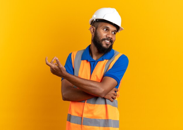 Homem jovem construtor afro-americano satisfeito de uniforme com capacete de segurança, olhando para o lado isolado em um fundo laranja com espaço de cópia