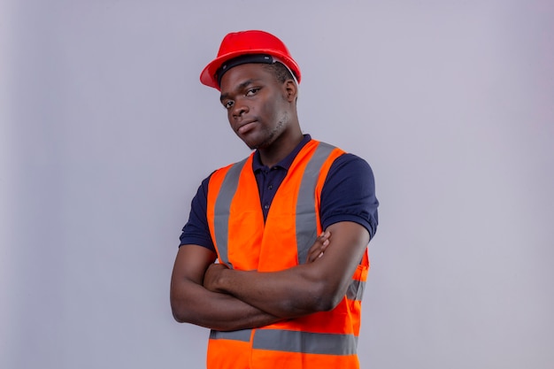 Homem jovem construtor afro-americano usando colete de construção e capacete de segurança em pé com os braços cruzados, parecendo suspeito