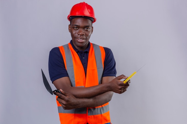 Foto grátis homem jovem construtor afro-americano usando colete de construção e capacete de segurança em pé com os braços cruzados segurando uma espátula parecendo confiante