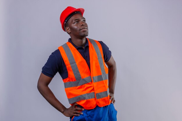 Homem jovem construtor afro-americano usando colete de construção e capacete de segurança, olhando para cima com o rosto sério em pé