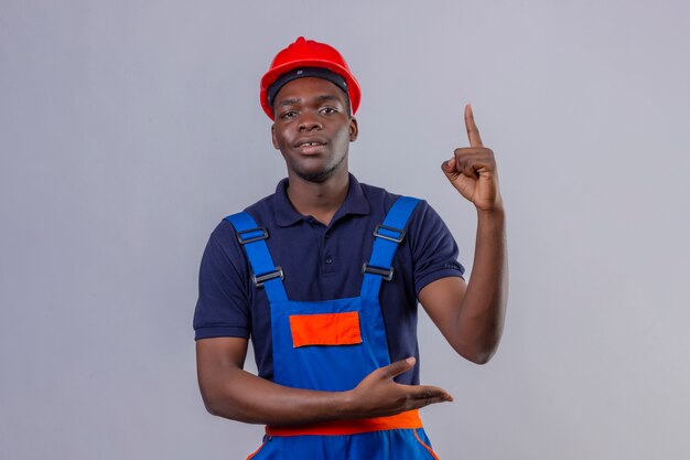Homem jovem construtor afro-americano usando uniforme de construção e capacete de segurança apontando o dedo para cima e apresentando-se com a palma da mão com expressão confiante em pé