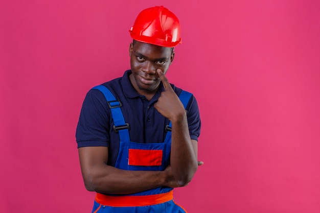 Homem jovem construtor afro-americano usando uniforme de construção e capacete de segurança apontando para o olho, vendo você fazer um gesto de expressão suspeita em pé no rosa isolado