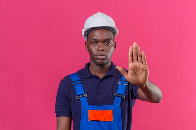 Homem jovem construtor afro-americano usando uniforme de construção e capacete de segurança em pé com a mão aberta fazendo sinal de pare com expressão séria e confiante gesto de defesa em pé
