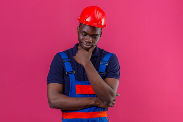 Homem jovem construtor afro-americano usando uniforme de construção e capacete de segurança em pé com a mão no queixo pensando tentando fazer uma escolha tendo dúvidas sobre rosa isolado