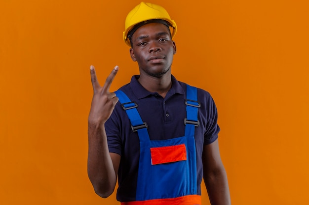 Homem jovem construtor afro-americano usando uniforme de construção e capacete de segurança, parecendo confiante, mostrando a vitória cantando em pé na laranja