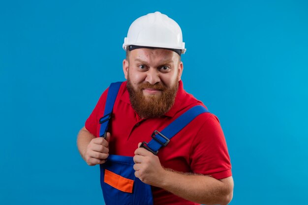 Homem jovem construtor barbudo com uniforme de construção e capacete de segurança descontente com a cara zangada