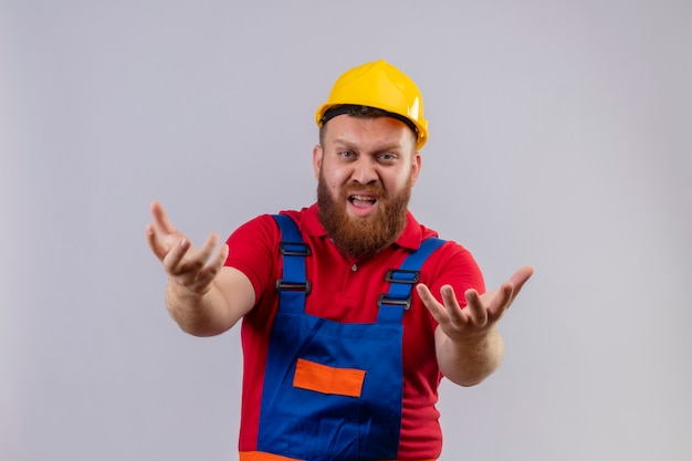 Homem jovem construtor barbudo com uniforme de construção e capacete de segurança, irritado e frustrado, gritando com os braços levantados