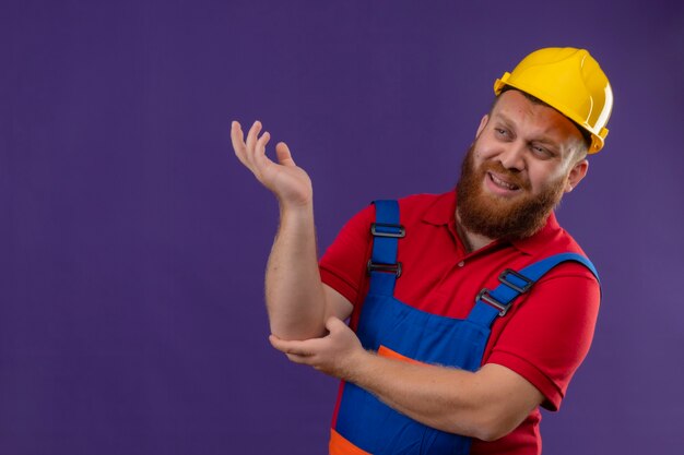 Homem jovem construtor barbudo com uniforme de construção e capacete de segurança, sorrindo alegremente, levantando o braço sobre o fundo roxo