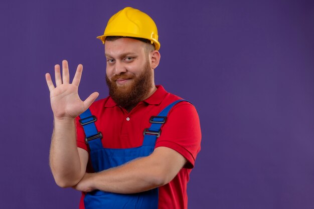 Homem jovem construtor barbudo com uniforme de construção e capacete de segurança sorrindo, mostrando e apontando para cima com os dedos número cinco sobre o fundo roxo