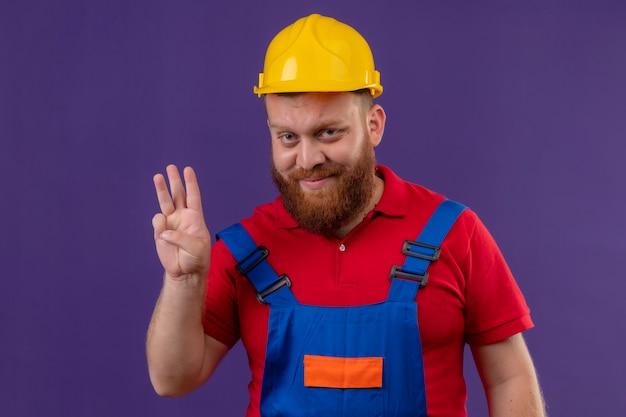 Homem jovem construtor barbudo com uniforme de construção e capacete de segurança sorrindo, mostrando e apontando para cima com os dedos número três sobre o fundo roxo