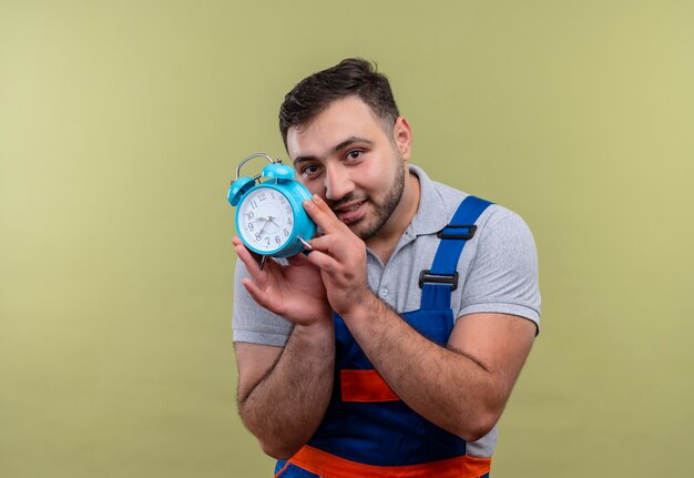 Homem jovem construtor com uniforme de construção segurando um despertador e olhando para a câmera com astúcia