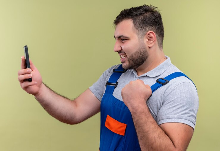 Foto grátis homem jovem construtor com uniforme de construção segurando um smartphone olhando para uma tela se passando por um vencedor com o punho cerrado feliz e animado