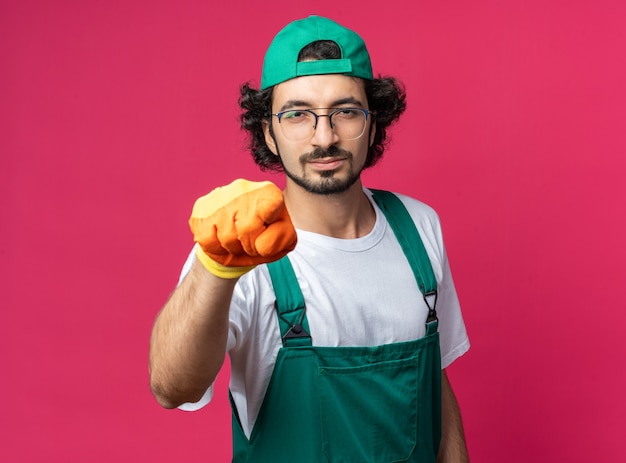 Homem jovem construtor confiante vestindo uniforme com boné e pontas de luvas na frente