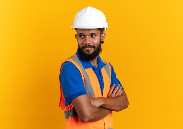 homem jovem construtor de uniforme com capacete de segurança em pé com os braços cruzados, isolado na parede laranja com espaço de cópia