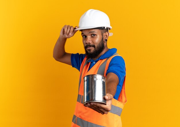 Homem jovem construtor de uniforme com capacete de segurança segurando tinta a óleo isolada na parede laranja com espaço de cópia