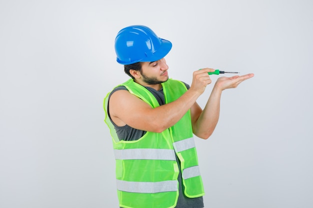 Homem jovem construtor de uniforme usando a chave de fenda enquanto trabalhava e olhava com foco, vista frontal.