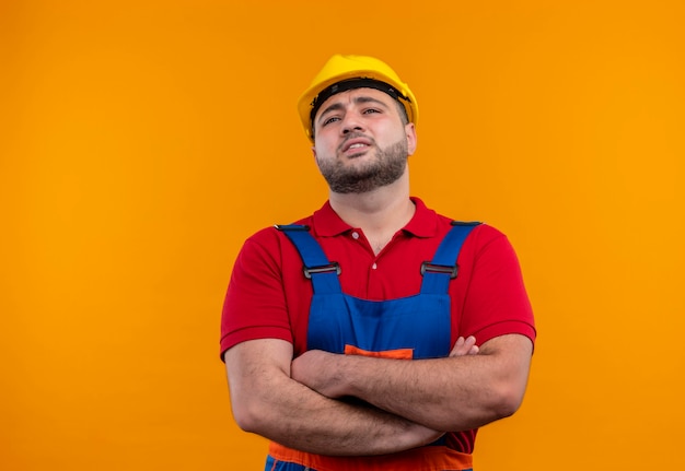 Homem jovem construtor descontente com uniforme de construção e capacete de segurança com as mãos cruzadas no peito