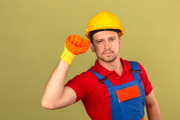 Homem jovem construtor em luvas de uniforme de construção e capacete de segurança, levantando o punho com o conceito de vencedor de rosto sério sobre parede verde isolada