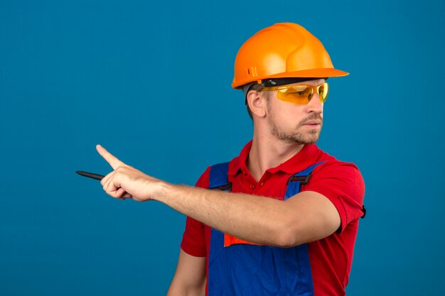 Homem jovem Construtor em uniforme de construção e capacete de segurança, apontando com o dedo indicador para o lado ao longo da parede azul isolada