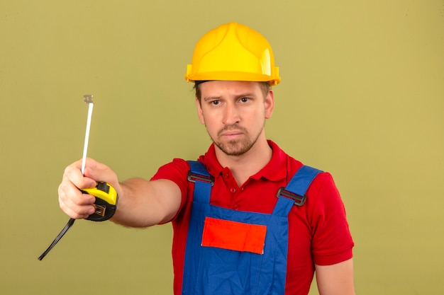 Foto grátis homem jovem construtor em uniforme de construção e capacete de segurança, dando a alguém fita-line com cara séria sobre parede verde isolada