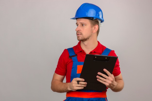 Foto grátis homem jovem construtor em uniforme de construção e capacete de segurança em pé com a área de transferência na mão e desviar o olhar com cara séria sobre parede branca isolada