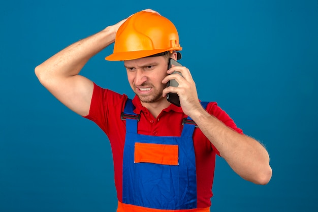 Homem jovem construtor em uniforme de construção e capacete de segurança, falando no telefone móvel infeliz em estresse ao longo da parede isolada azul