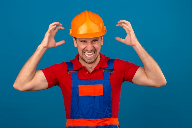 Homem jovem construtor em uniforme de construção e capacete de segurança louco gritando e gritando com expressão agressiva e braços levantados sobre parede azul isolada