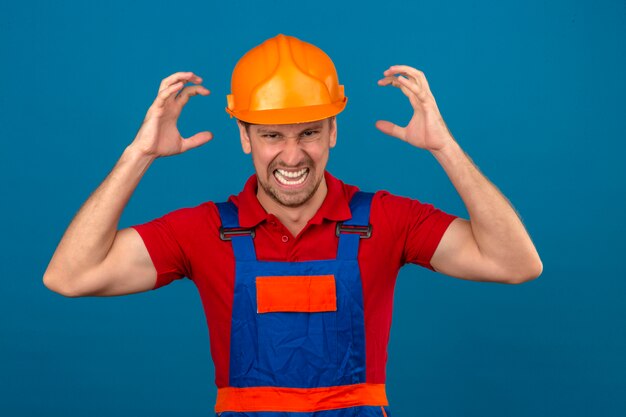 Homem jovem Construtor em uniforme de construção e capacete de segurança louco gritando e gritando com expressão agressiva e braços levantados sobre parede azul isolada