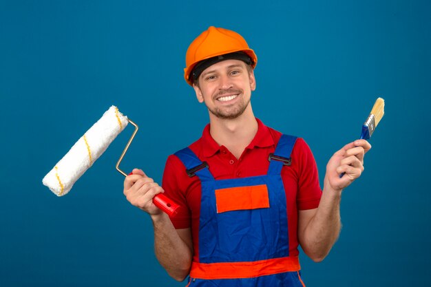 Homem jovem Construtor em uniforme de construção e capacete de segurança, segurando o rolo de pintura e pincel com um grande sorriso no rosto sobre parede azul isolada