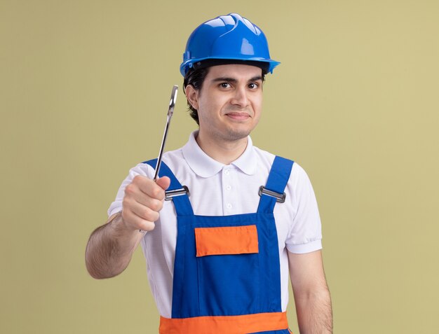 Homem jovem construtor em uniforme de construção e capacete de segurança segurando uma chave inglesa, olhando para a frente, sorrindo em pé sobre a parede verde