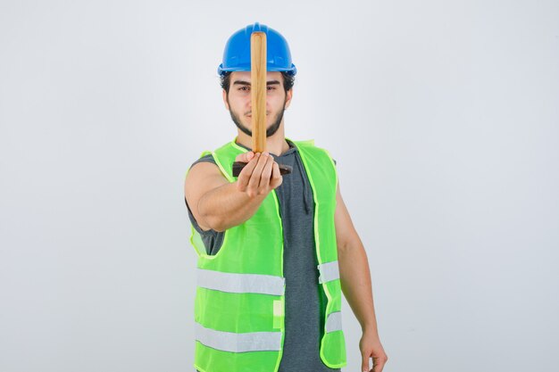 Homem jovem construtor em uniforme de trabalho, arranhando o martelo em direção à câmera e parecendo autoconfiante, vista frontal.