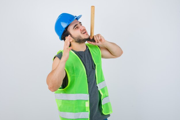 Homem jovem construtor em uniforme de trabalho, segurando o martelo enquanto levanta a mão e parece alegre, vista frontal.