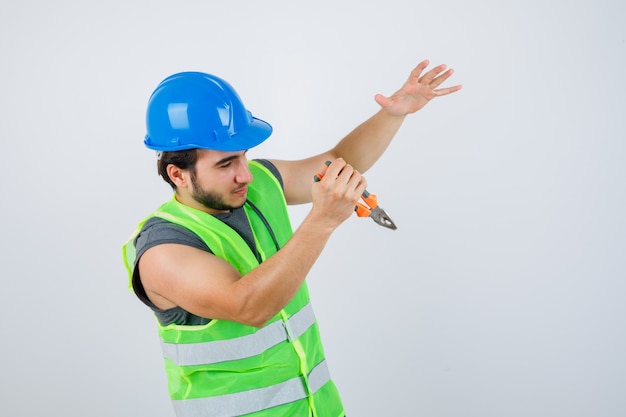 Homem jovem construtor em uniforme de trabalho, segurando um alicate enquanto finge pegar algo e parece autoconfiante, vista frontal.