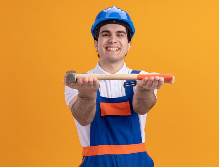 Foto grátis homem jovem construtor feliz com uniforme de construção e capacete de segurança, demonstrando o martelo olhando para a frente, sorrindo alegremente em pé sobre a parede laranja