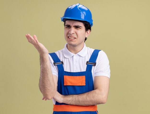Homem jovem construtor insatisfeito com uniforme de construção e capacete de segurança, olhando para o lado, levantando a mão com desprazer e indignação em pé sobre a parede verde