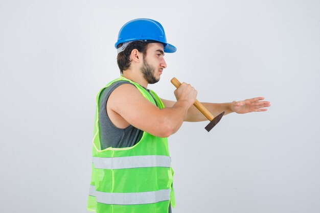 Homem jovem construtor, rememorando algo com o uso de um martelo em uniforme de trabalho e parecendo confiante, vista frontal.